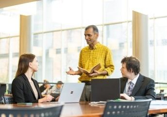 A man standing near two people seated at laptops, engaged in conversation
